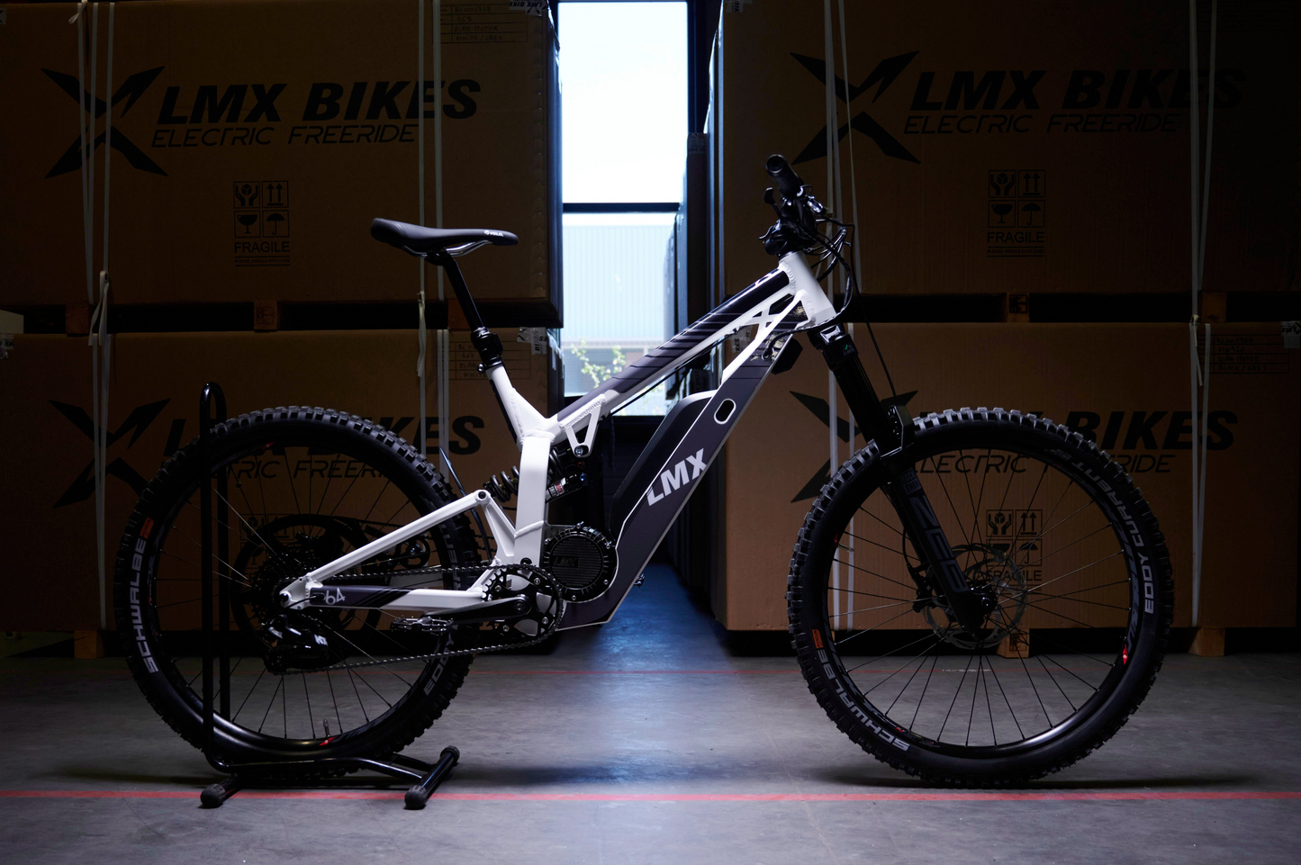 An LMX - 64 electric mountain bike featuring a mid-mount motor in black and white is showcased in a warehouse, surrounded by large boxes branded "LMX Bikes Electric Freeride.