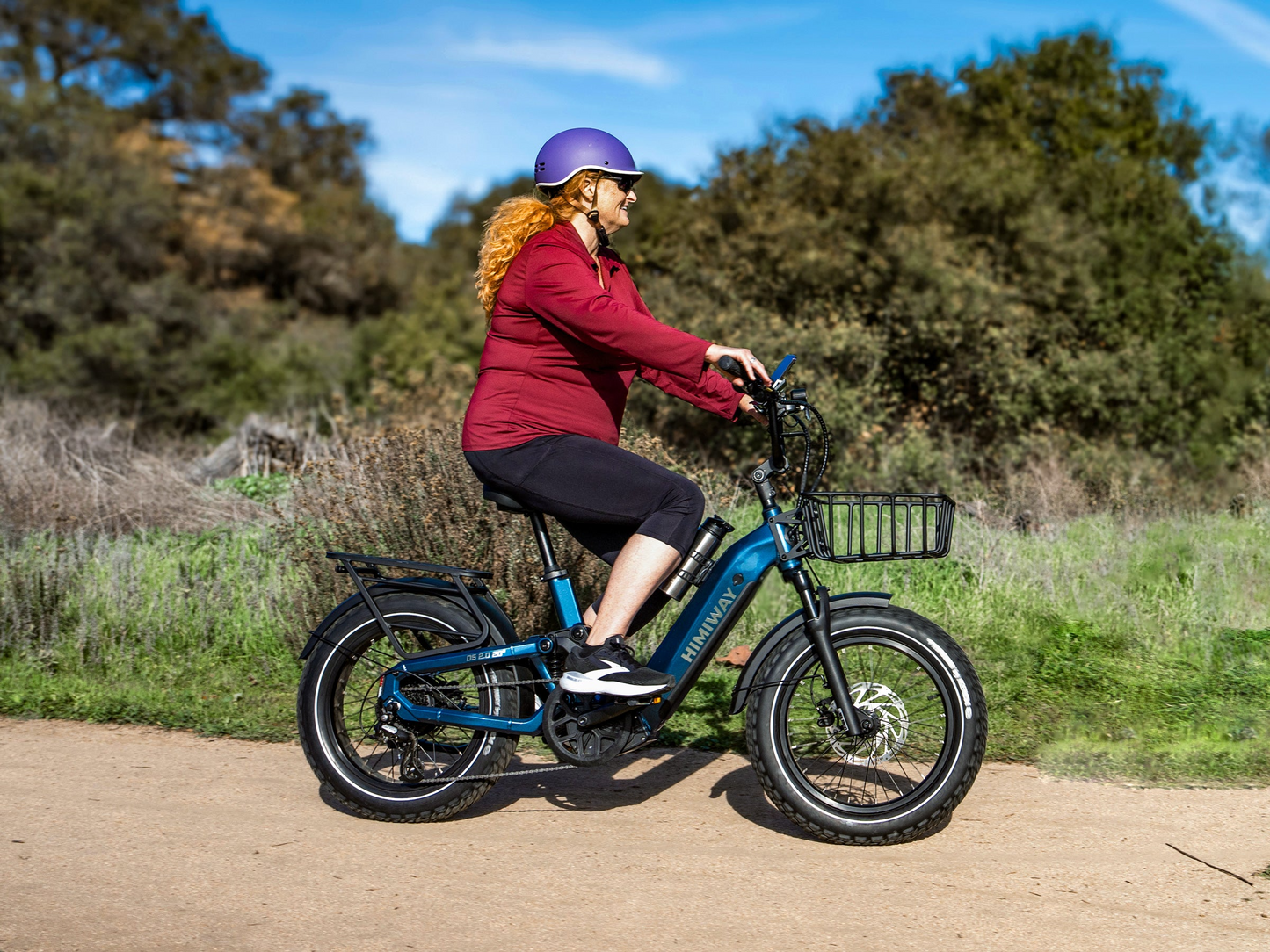 A person in a purple helmet and red jacket rides the Himiway D5 2.0 20in full-suspension eBike with fat tires on a dirt path, surrounded by greenery and trees.