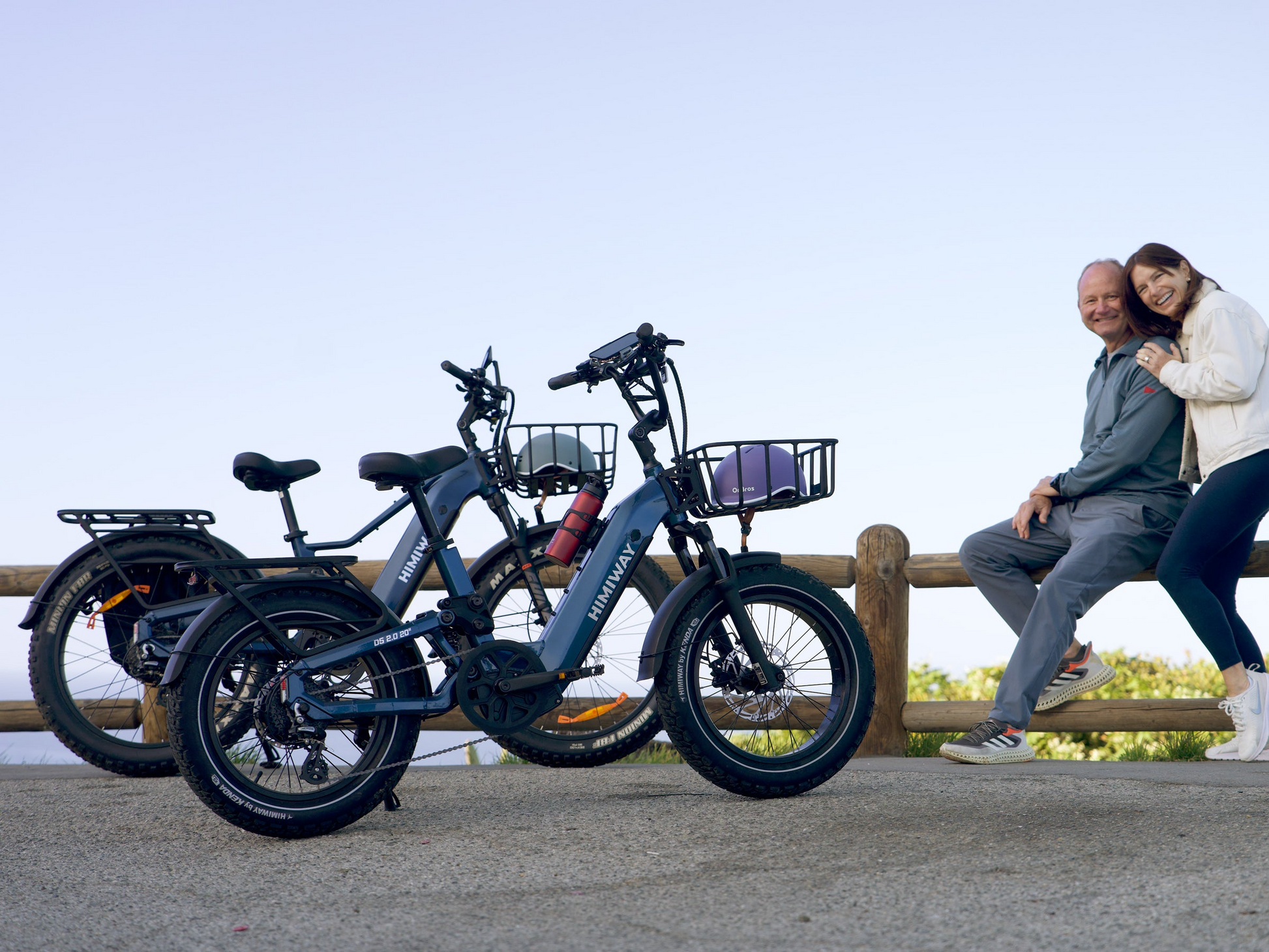 Two Himiway D5 2.0 20in fat tire electric bikes by Himiway are parked on pavement near a wooden fence as an older man and young woman smile beside them, each featuring a powerful 750W hub motor under a clear sky.