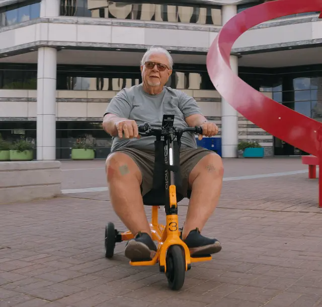 An older man rides a yellow Dashmoto Dash 3 folding mobility scooter by Dashmoto outside a modern building, with a striking red spiral sculpture in the background.