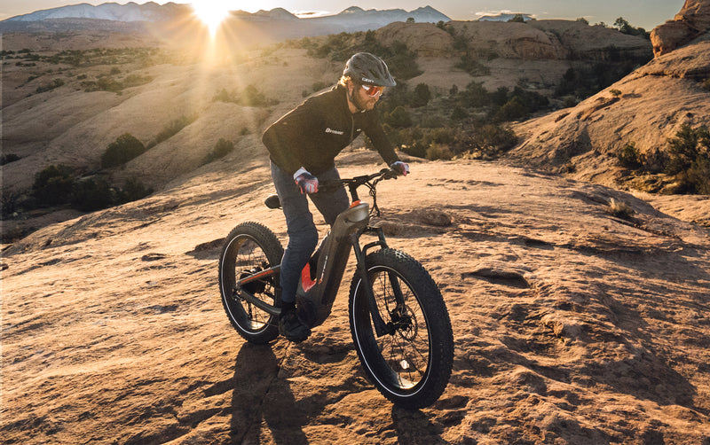 A person biking on rocky terrain during sunset; the landscape features smooth, arid rock formations with a low sun in the background while riding a HeyBike - Hero (Mid-Drive).