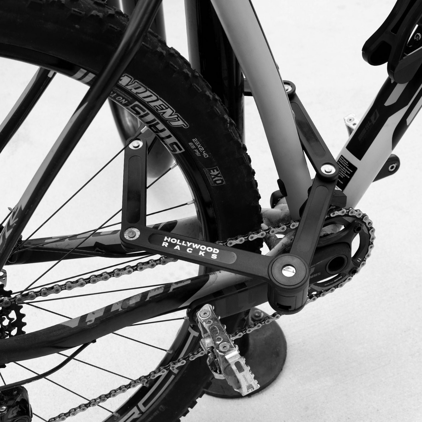 A close-up black and white image of a bicycle secured to a bike rack. The bike features a "Hollywood Racks" brand mount and a Hollywood Racks Lock - Folding attached near the rear wheel and pedal, enhancing its high security.