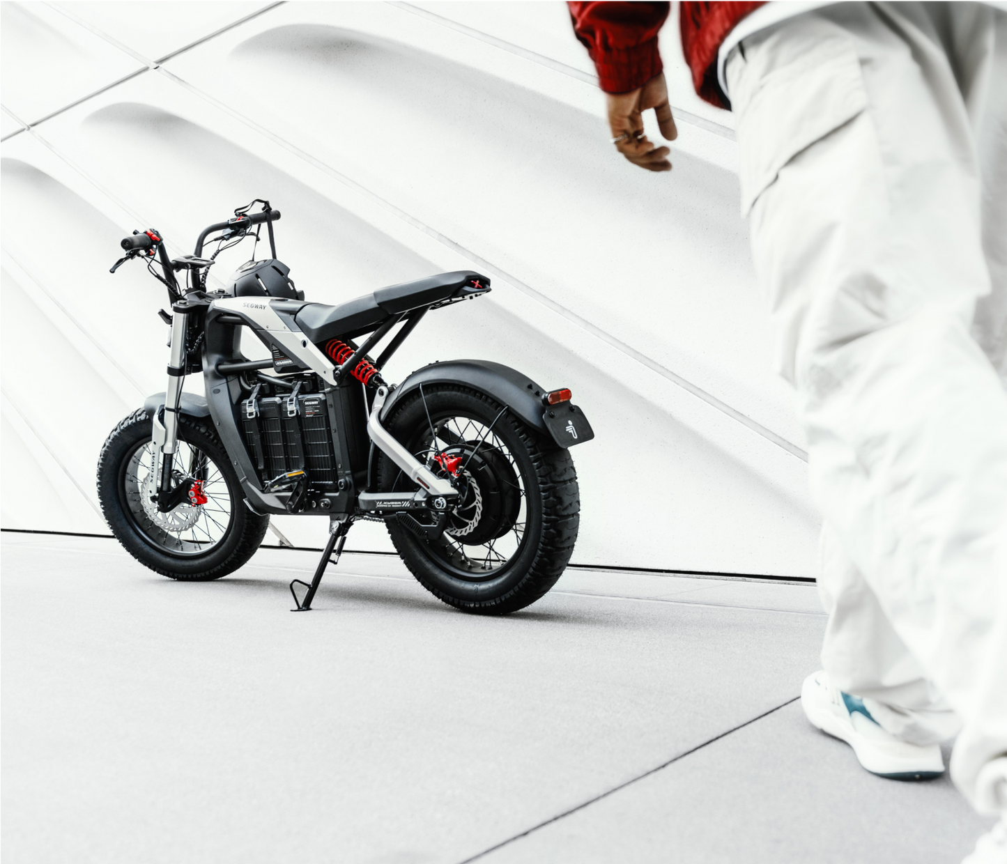 A stylish Segway - Xyber, featuring a black frame with red accents, is poised on the pavement. A person in white pants and a red jacket walks by, against a bold white striped backdrop, embodying Tampa Bay's vibrant energy.