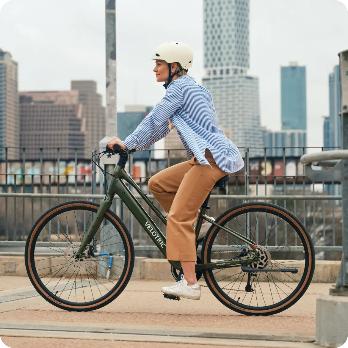 Wearing a helmet, striped shirt, and tan pants, a person rides the Velotric Tempo eBike by Velotric along an urban path with city buildings in the background.