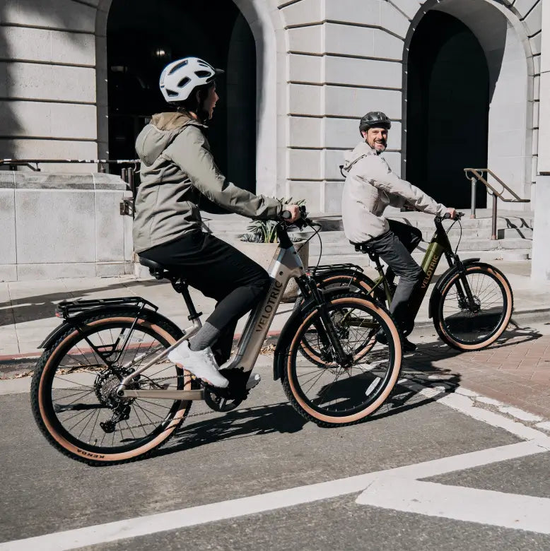 Two people wearing helmets ride Velotric Discover M eBikes by Velotric along a city street, passing a building with arched doorways.