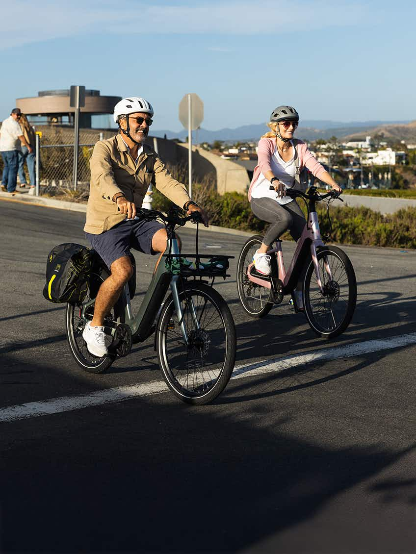 Two adults in helmets ride Aventon Pace 5 REC Step-Through electric bikes by Aventon on a paved coastal road, with hills and buildings in the background.