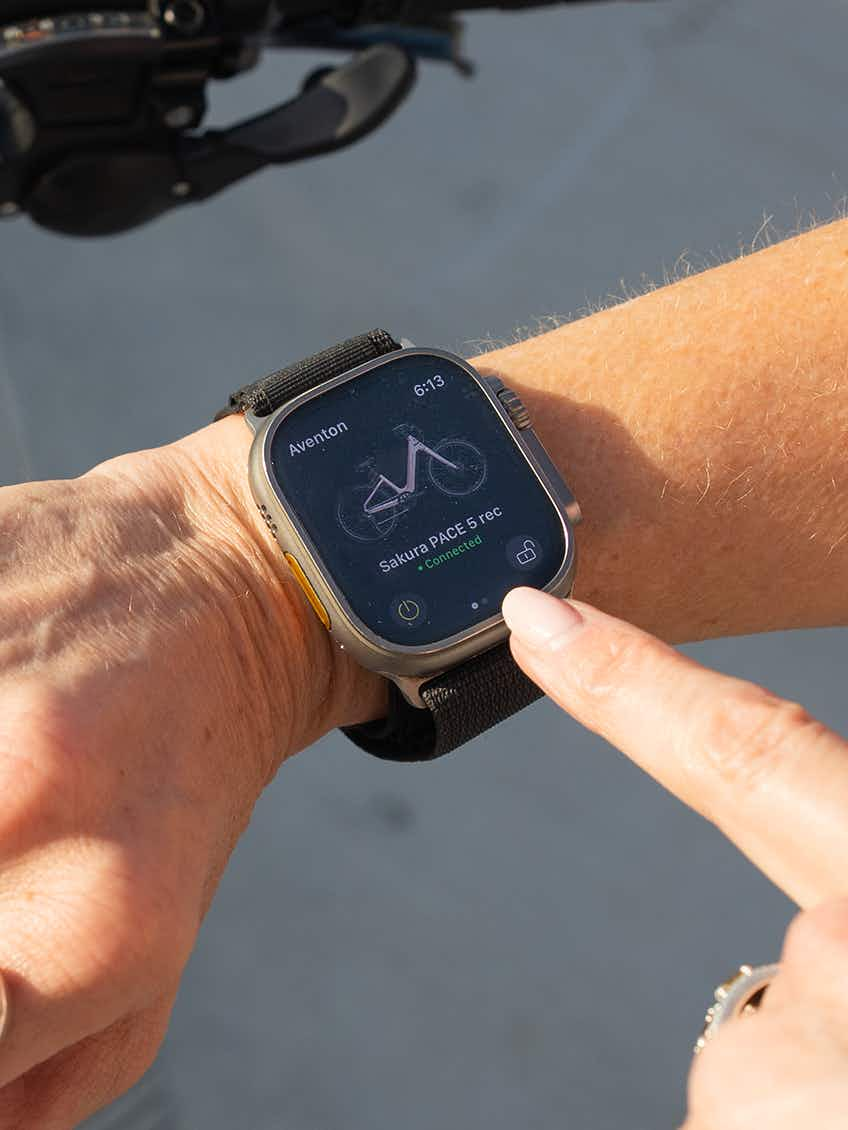 A hand interacts with a smartwatch showing a cycling activity tracker while riding an Aventon Pace 5 REC Step-Through electric bike by Aventon.