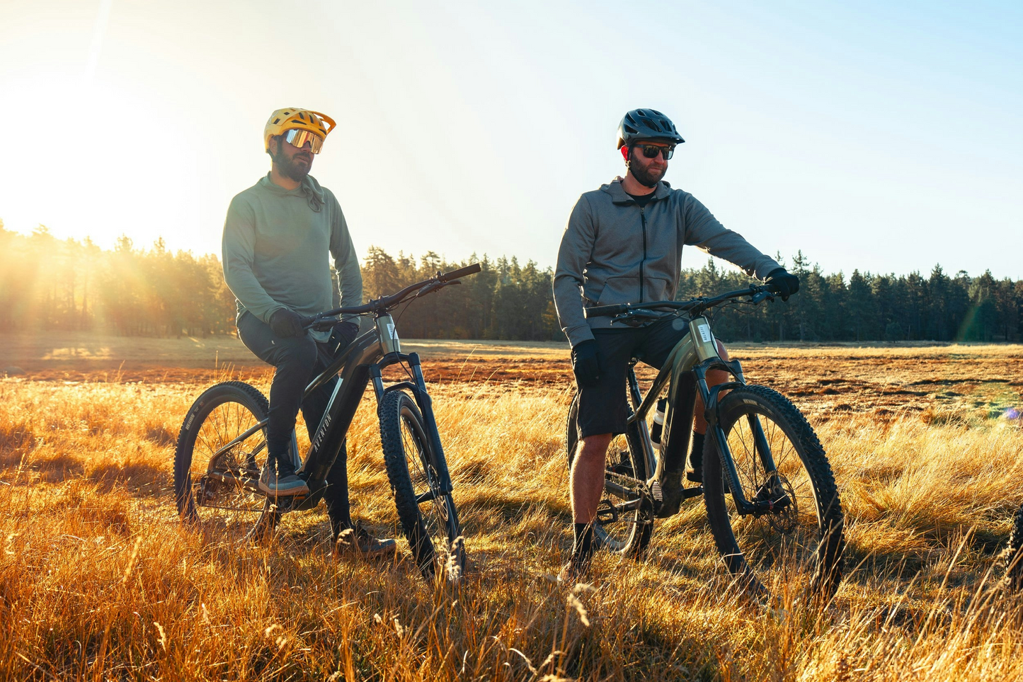 Two cyclists with Aventon Ramblas eMTBs standing in a field at sunrise.