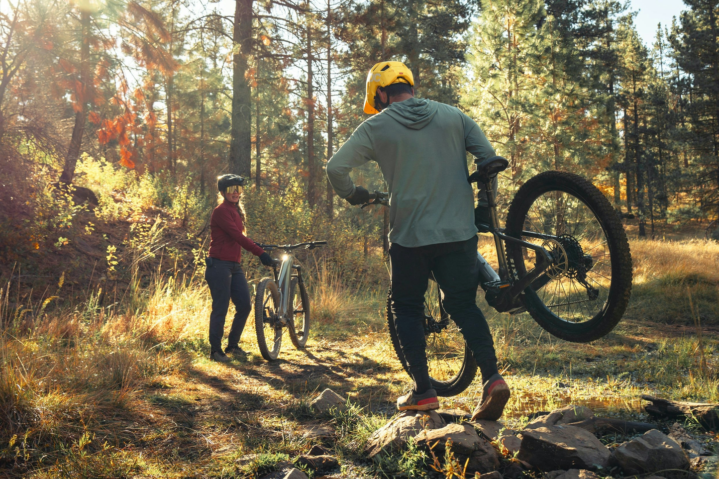 Two mountain bikers having a conversation in a sunny forest clearing, one of them leaning on their Aventon Ramblas eMTB electric mountain bike.