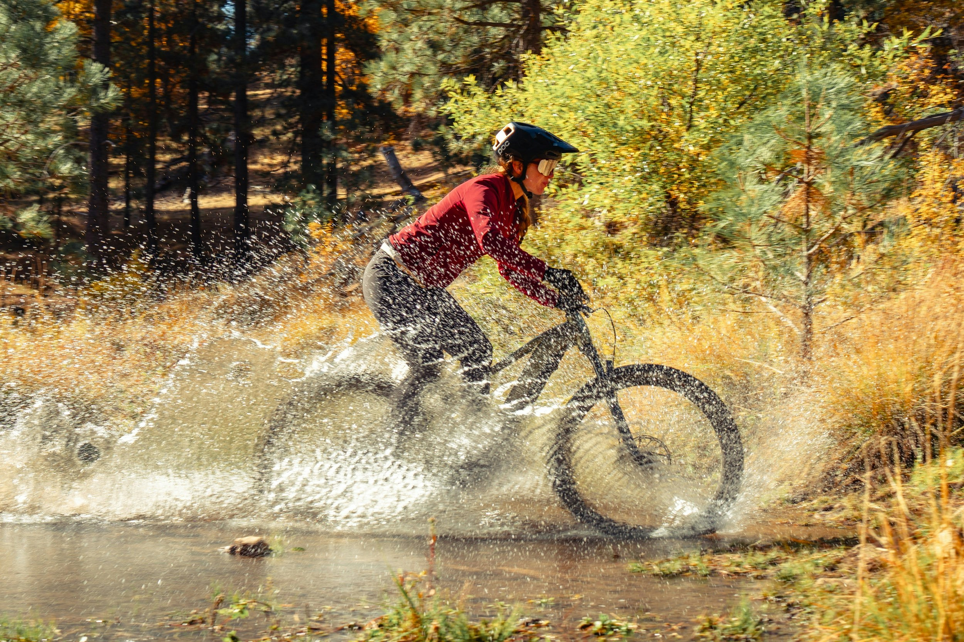 A mountain biker riding an Aventon Ramblas eMTB through a shallow stream, creating a splash.