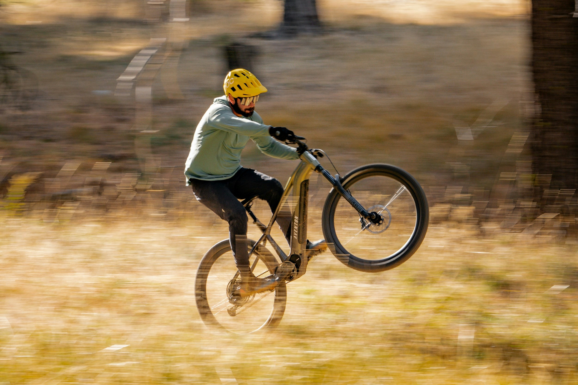 A cyclist in motion, performing a wheelie on an Aventon - Ramblas eMTB electric mountain bike on a dirt track while wearing a yellow helmet.