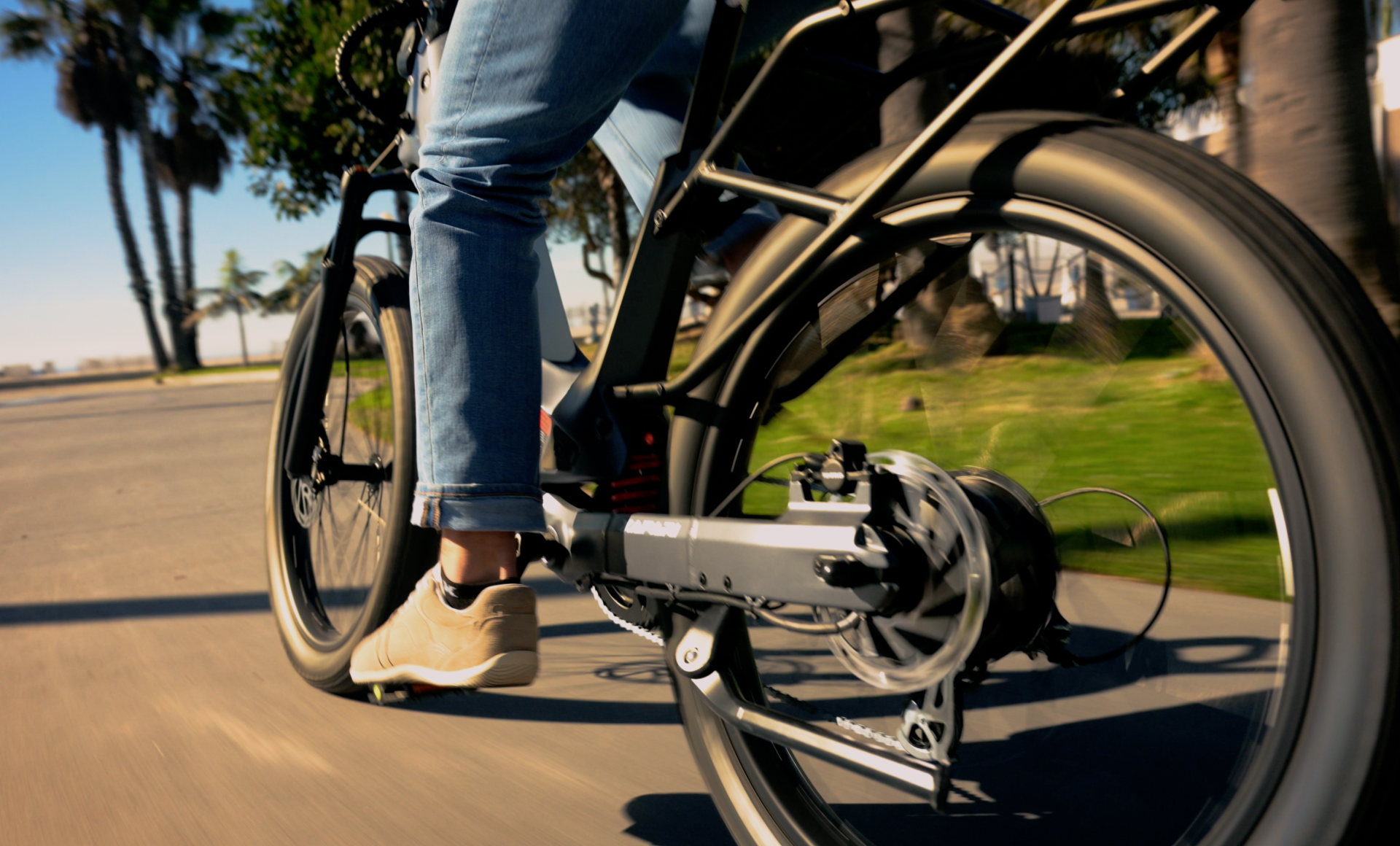 A close-up shows a person riding the Segway Xafari on a sunny path, with palm trees gently swaying nearby. The focus is on the bike's rear wheel and motor, effortlessly providing an extended range for adventurous trips.