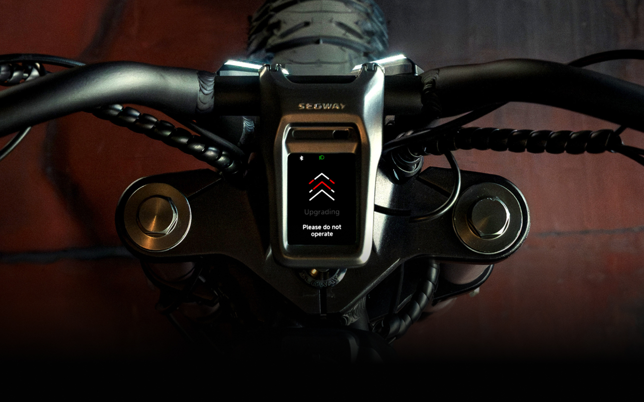 A close-up of a Segway - Xyber handlebar features a digital display with an "Upgrading" message and arrows, signaling not to operate.