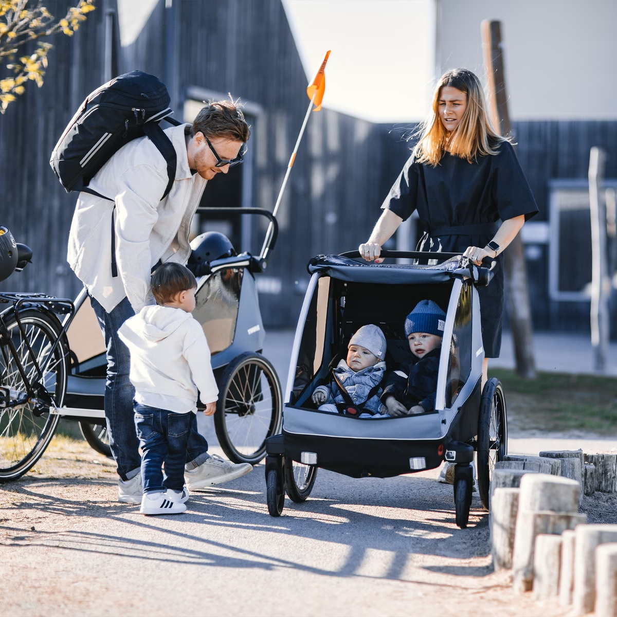 A man and woman are outdoors with three children. The man wears a backpack and talks to one child, while the woman pushes two children in a stroller. A Thule bicycle with the Thule Courier child trailer, perfect for family outings, stands nearby.