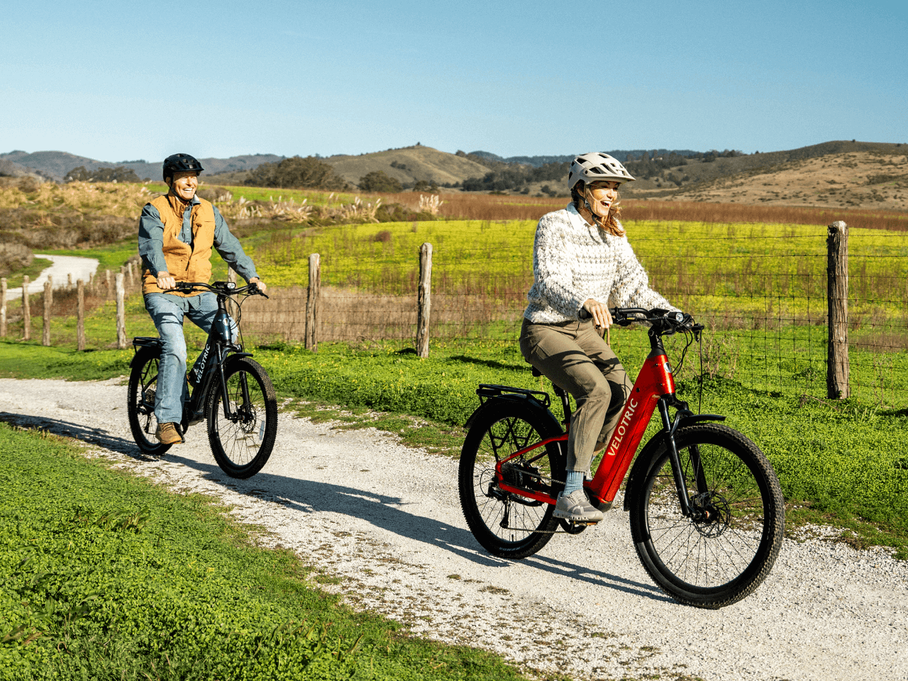 Two riders in helmets cruise on Velotric Discover 3 electric bikes by Velotric along a gravel path through grassy countryside beneath a clear Tampa Bay sky.