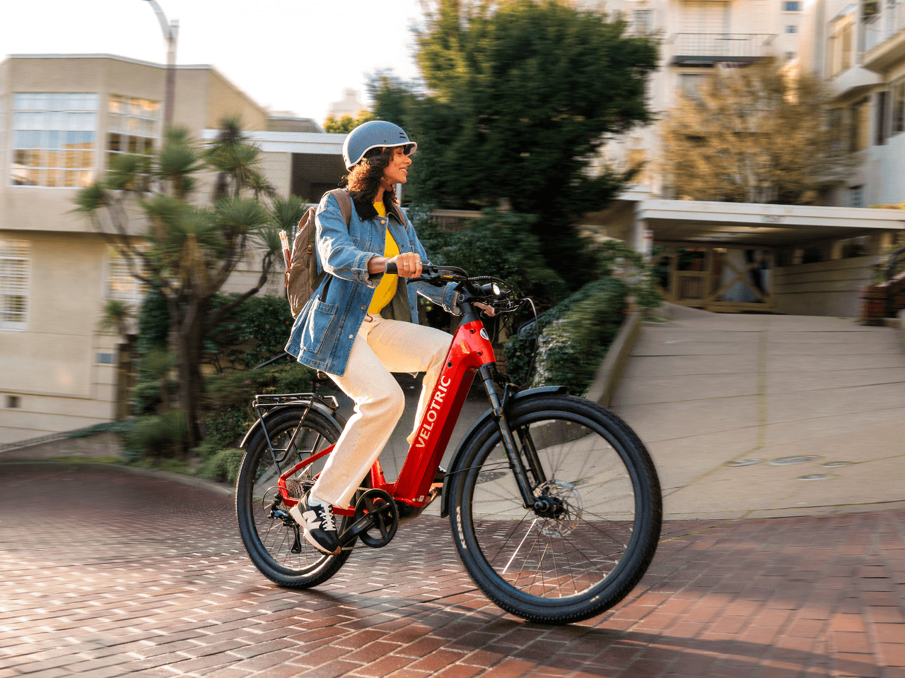 A person wearing a helmet and backpack rides a red Velotric Discover 3 electric bike by Velotric up a brick-paved street in an urban Tampa Bay neighborhood during the day.