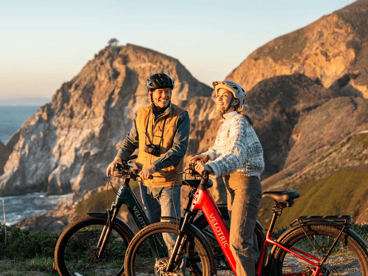 Two people in helmets stand with Velotric Discover 3 e-bikes from Velotric on a grassy hill above ocean cliffs near Tampa Bay, smiling at each other.