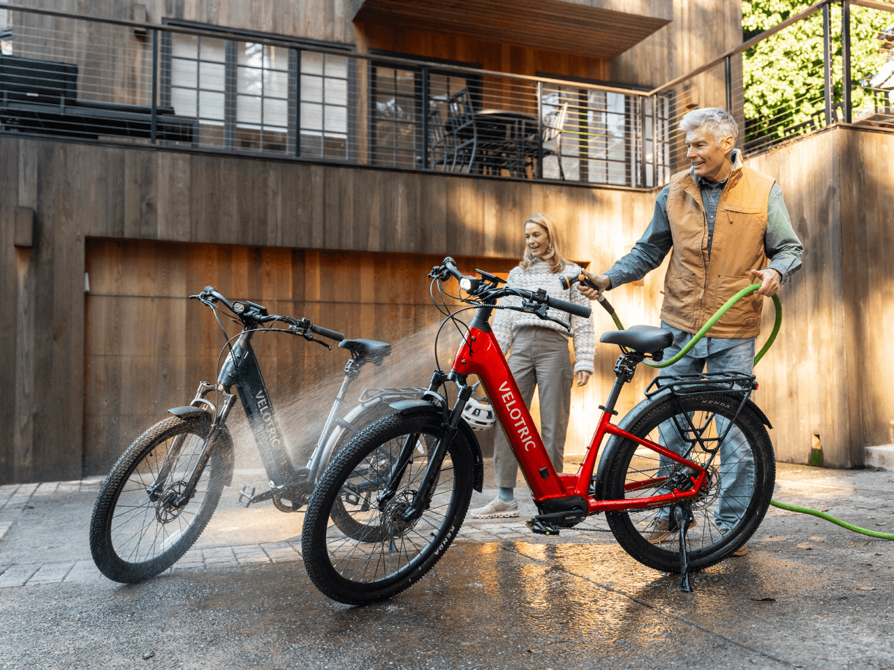 An older man sprays two bicycles—including the Velotric Discover 3 by Velotric—with a garden hose, while a woman stands nearby in front of a modern wooden building.