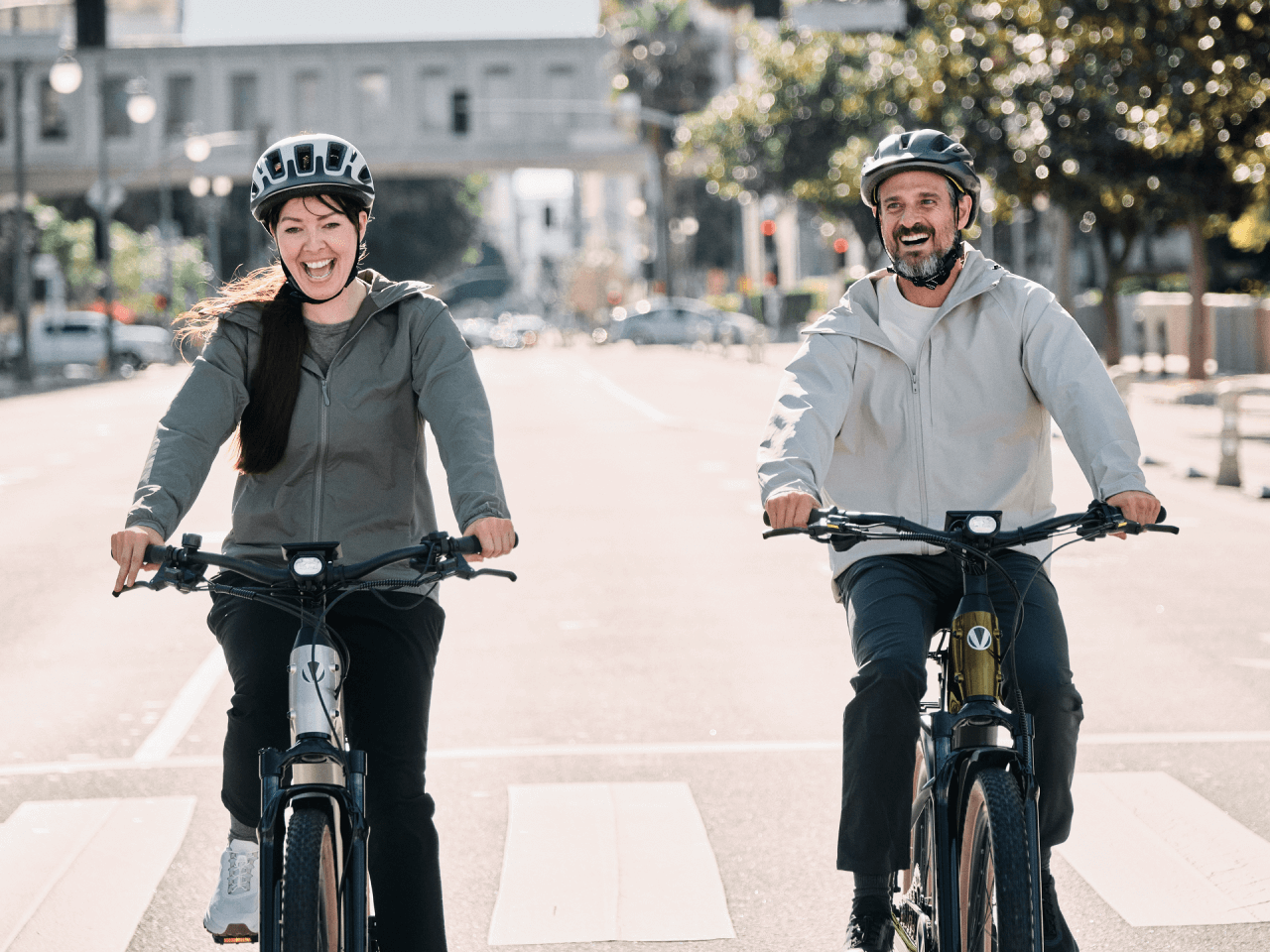 Two adults wearing helmets smile as they ride Velotric Discover M eBikes by Velotric side by side down a city street, framed by trees and buildings.