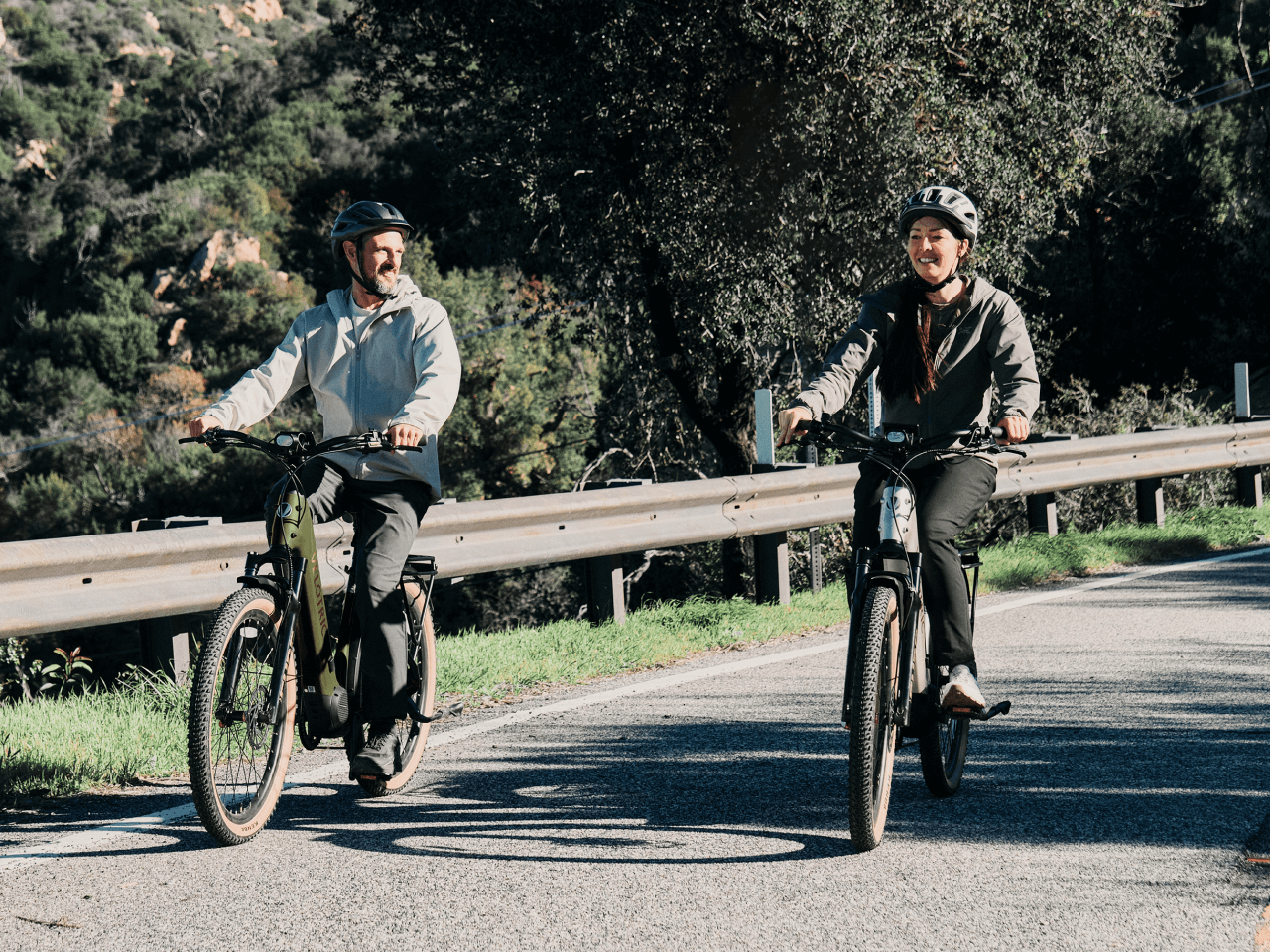 Two riders wearing helmets cruise side by side on Velotric Discover M eBikes by Velotric along a paved road, enjoying the smooth ride of this comfort commuter amid trees and a metal guardrail.