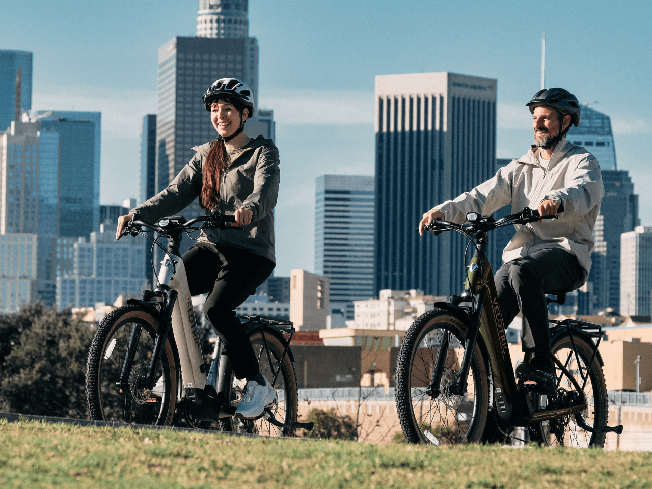 Two people, both in helmets, ride Velotric Discover M eBikes by Velotric across grassy terrain with tall city buildings in the background.