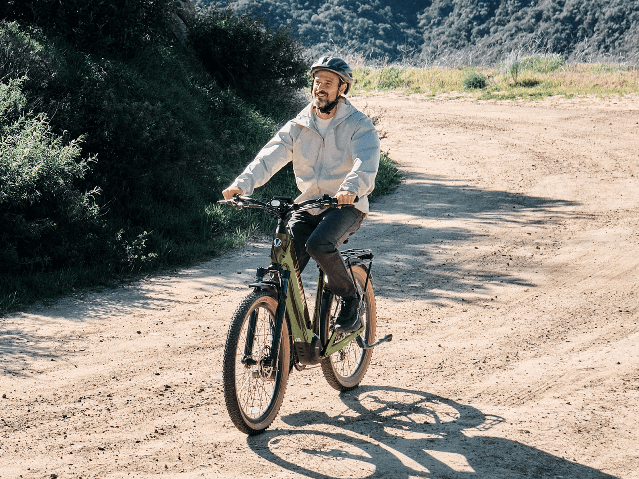 A man rides a Velotric Discover M eBike by Velotric along a dirt path with greenery and hills under a sunny sky.