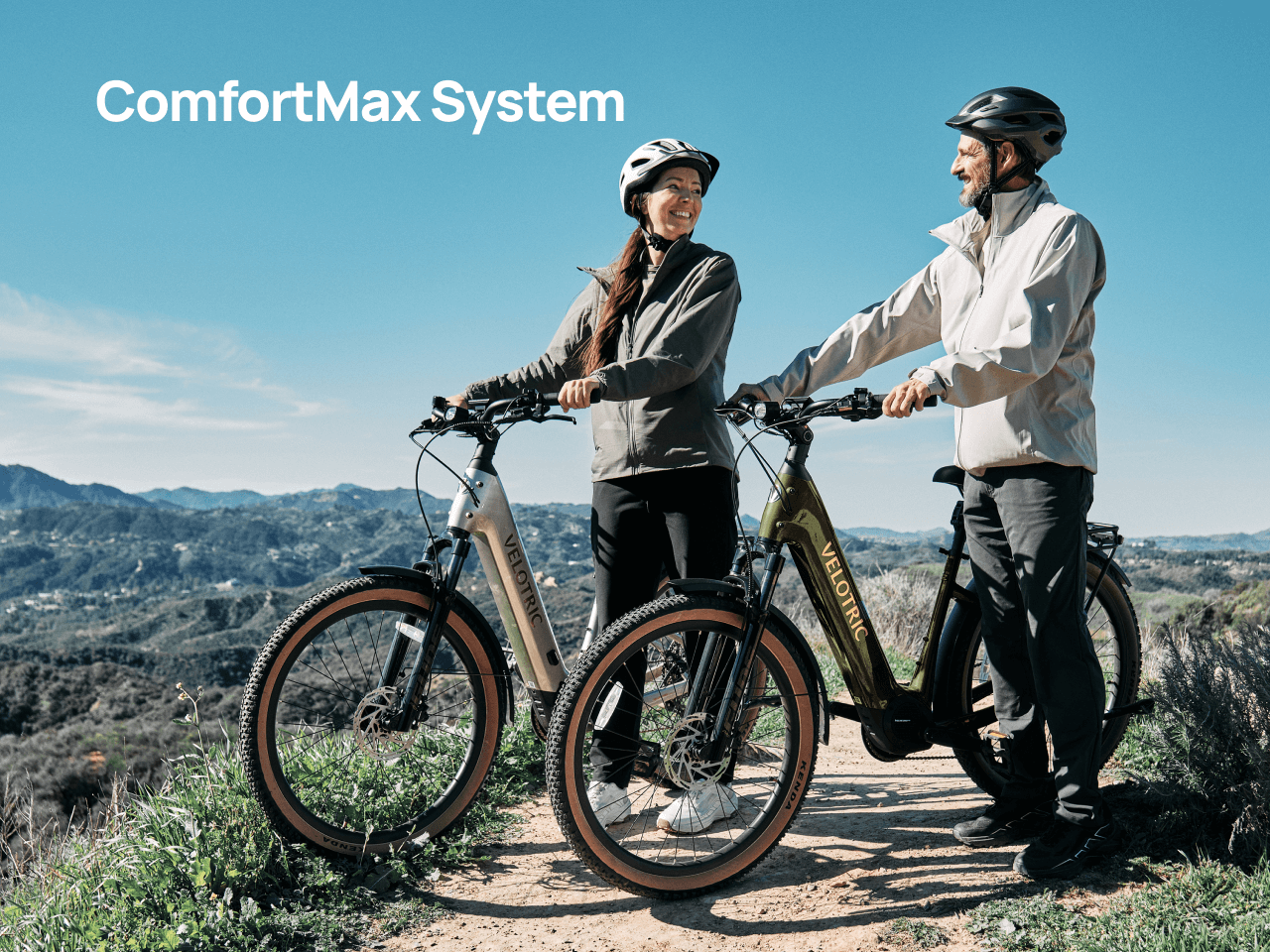 A woman and man in helmets smile at each other on a trail with Velotric Discover M eBikes; mountains and blue sky behind them. Text: "ComfortMax System." Brand: Velotric.