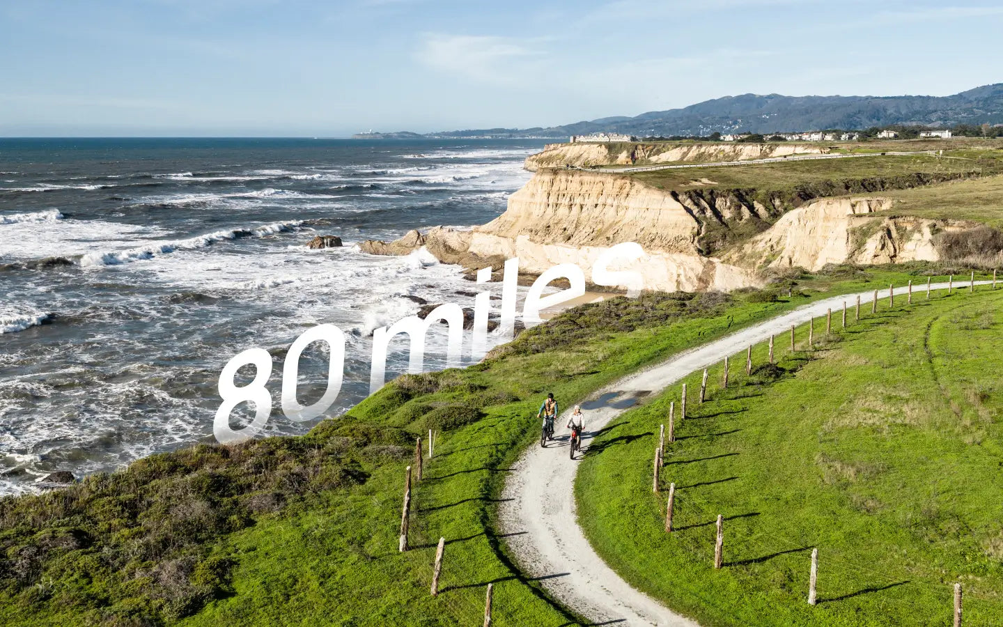 Aerial view of two cyclists riding Velotric Discover 3 eBikes by Velotric along a coastal trail near cliffs and the ocean, with "80 miles" displayed in large font.