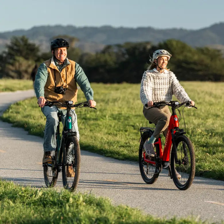 Two adults wearing helmets ride Velotric Discover 3 eBikes by Velotric along a paved path through grassy fields, with trees and hills in the background.