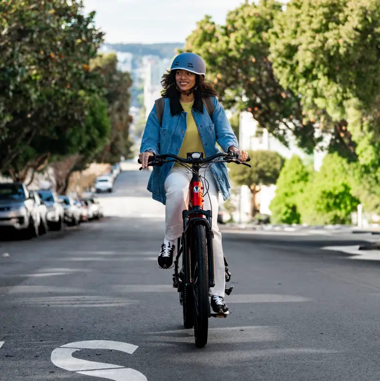 A person in a helmet and casual clothes rides the Velotric Discover 3 by Velotric along a quiet residential street with parked cars and trees.