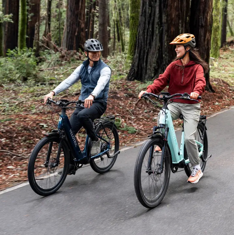 Two people wearing helmets ride Velotric Discover 3 eBikes by Velotric on a paved path through a forested area, smiling at each other.