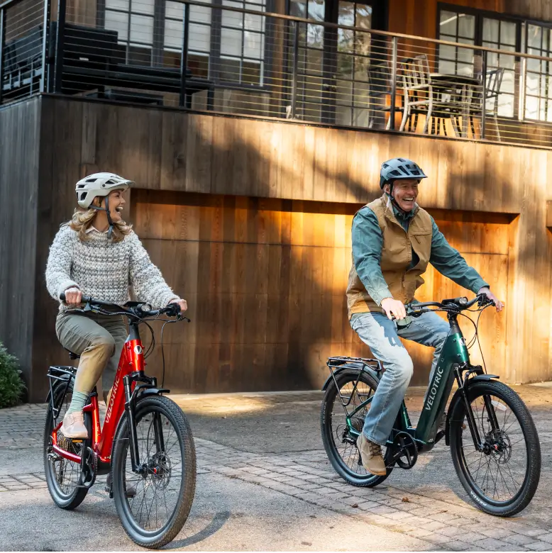 A man and a woman wearing helmets ride Velotric Discover 3 eBikes by Velotric on a paved path in front of a modern wooden building, highlighting a stylish way to explore Tampa Bay by electric bike.