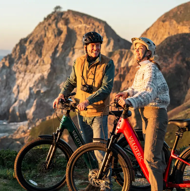 Two people wearing helmets smile at each other outdoors with the Velotric Discover 3 eBikes by Velotric, set against rocky cliffs and an ocean backdrop.