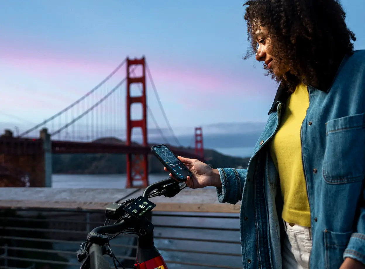 A woman stands beside her Velotric Discover 3 e-bike by Velotric, checking her smartphone with the Golden Gate Bridge silhouetted at sunset in the background.