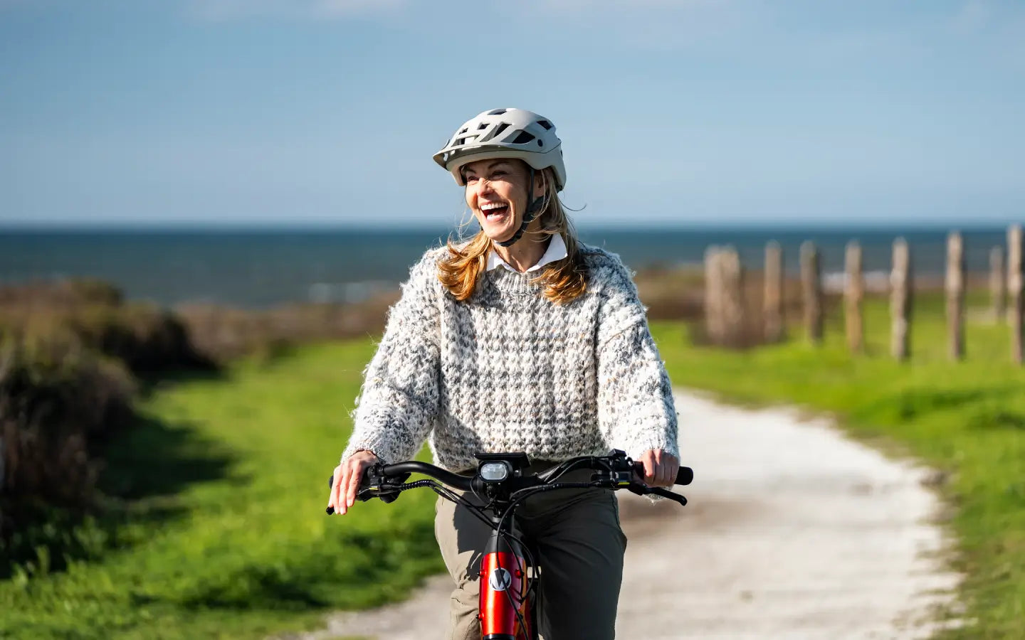 A woman wearing a helmet smiles as she rides the Velotric Discover 3 eBike by Velotric along a coastal gravel path, with green grass and the sea in the background.