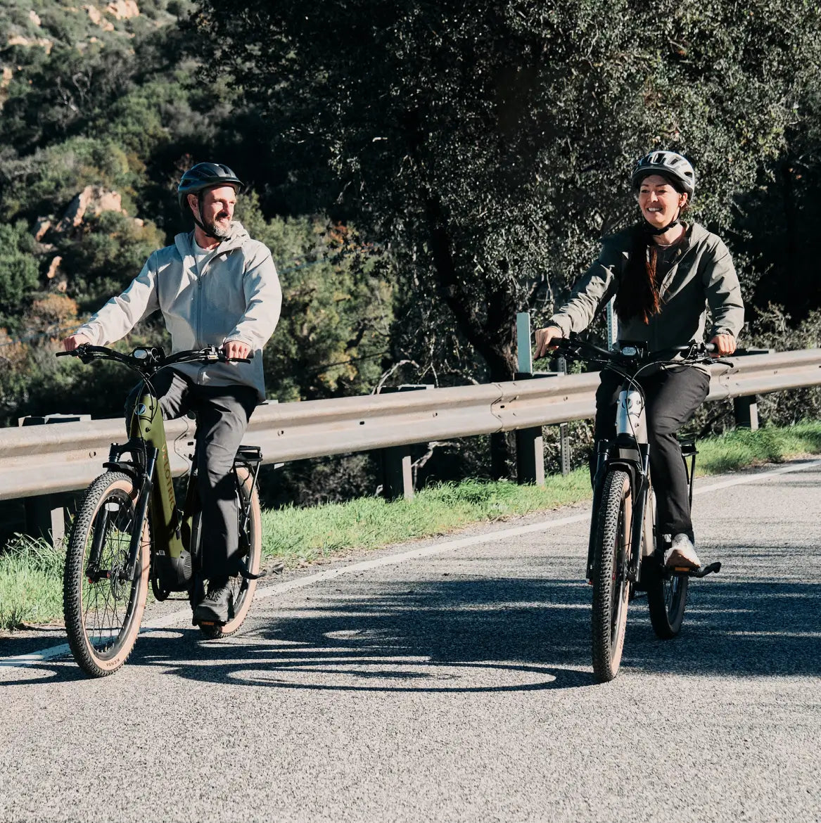 Two helmeted riders enjoy a Velotric Discover M eBike by Velotric on a paved road beside a guardrail, with trees and greenery in the background.