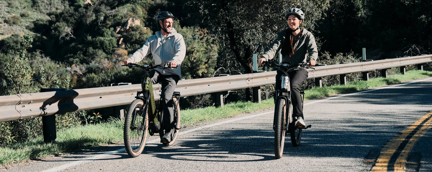 Two people wearing helmets ride Velotric Discover M eBikes by Velotric side by side on a paved road with guardrails, surrounded by trees and greenery.