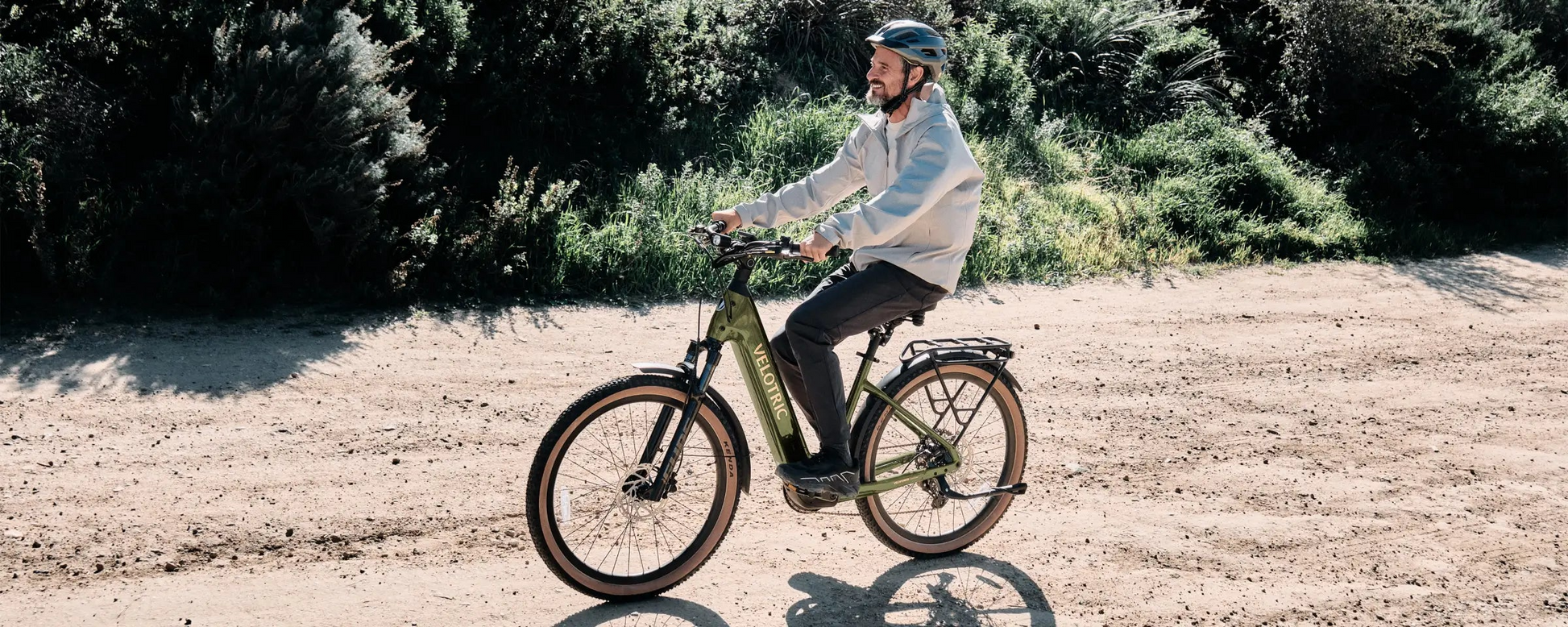 A man in a helmet rides the Velotric Discover M eBike by Velotric along a dirt path, surrounded by trees and shrubs.