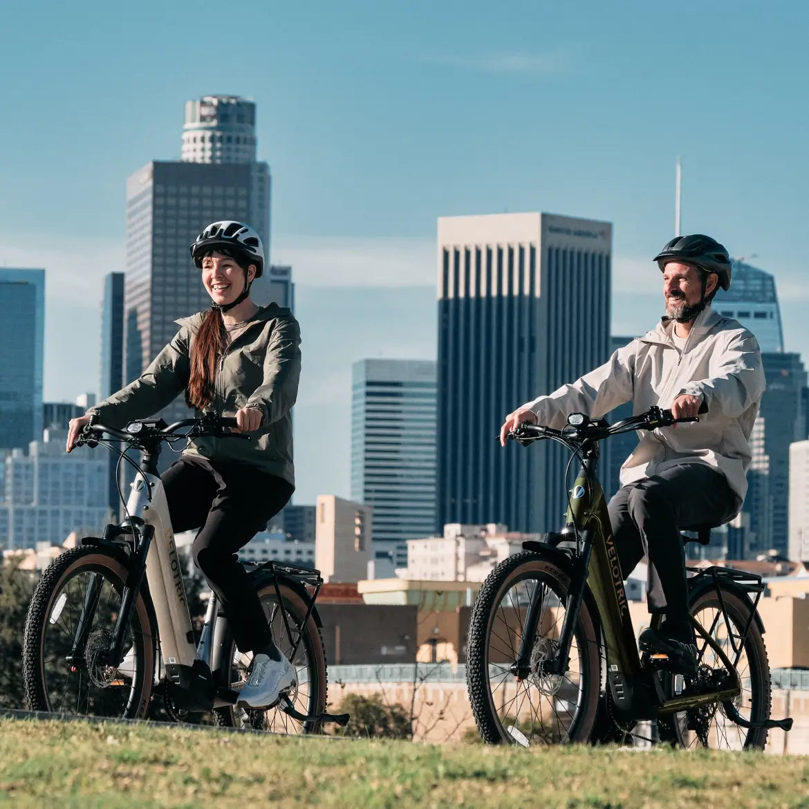 Two riders in helmets enjoy the Velotric Discover M, a comfort commuter eBike by Velotric, on grass with city skyscrapers behind them.