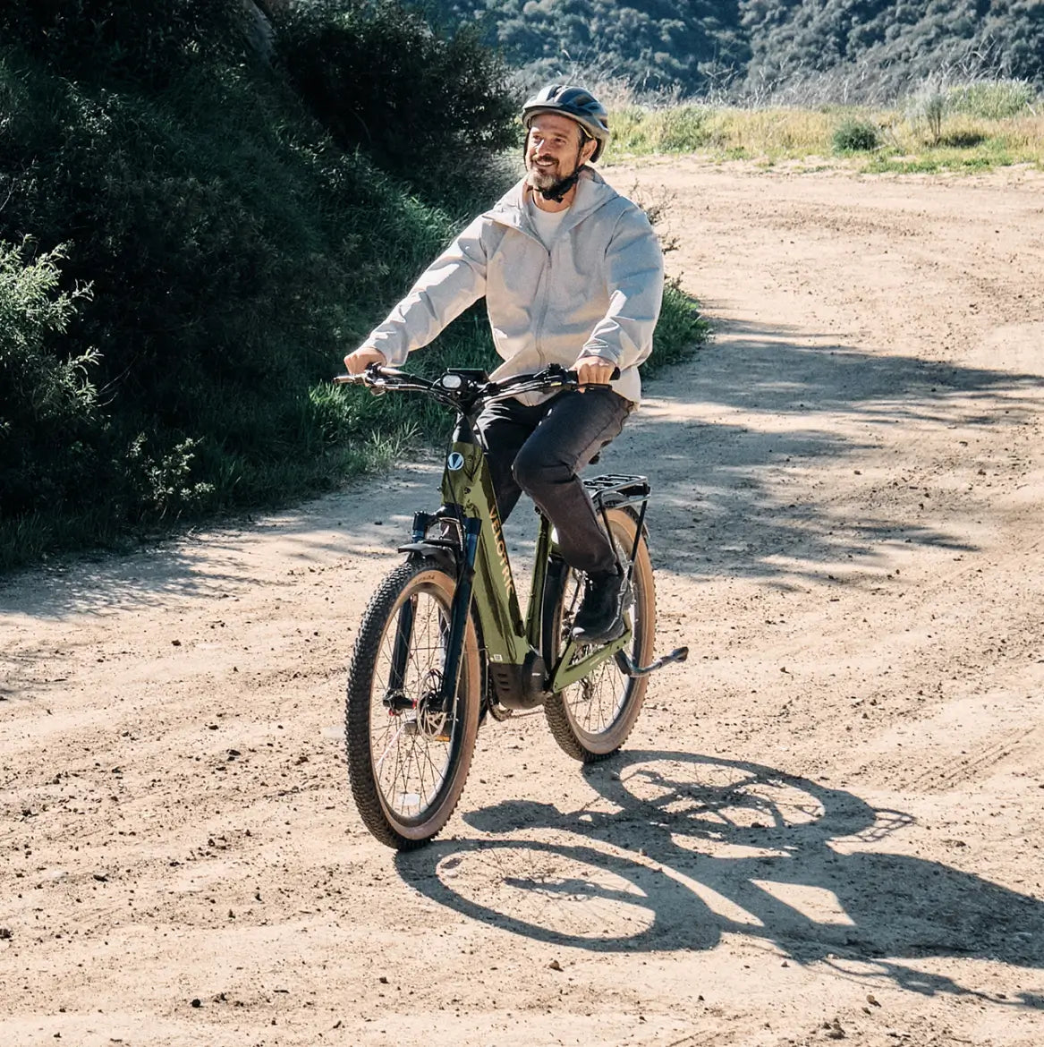 A person wearing a helmet and light jacket rides the Velotric Discover M commuter eBike by Velotric along a dirt trail with greenery and hills in the background.