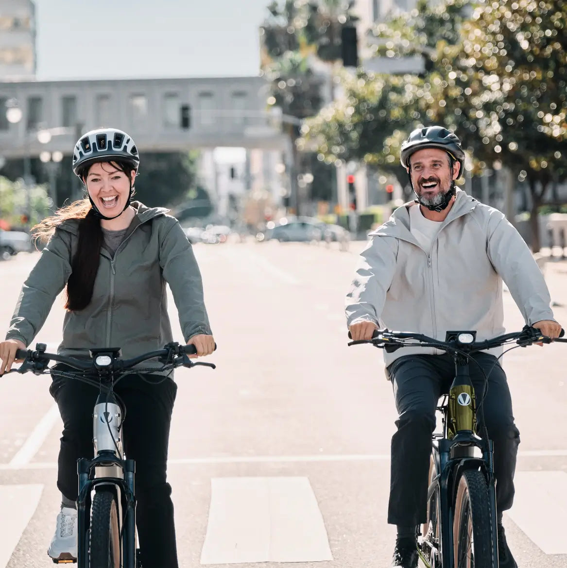Two adults in helmets and jackets ride Velotric Discover M eBikes from Velotric side by side on a city street, smiling as they cross a crosswalk in daylight.