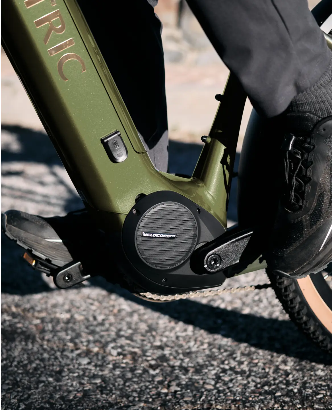Close-up of a person in black shoes riding the Velotric Discover M by Velotric, a comfort commuter eBike with a green frame, on a sunlit gravel path.
