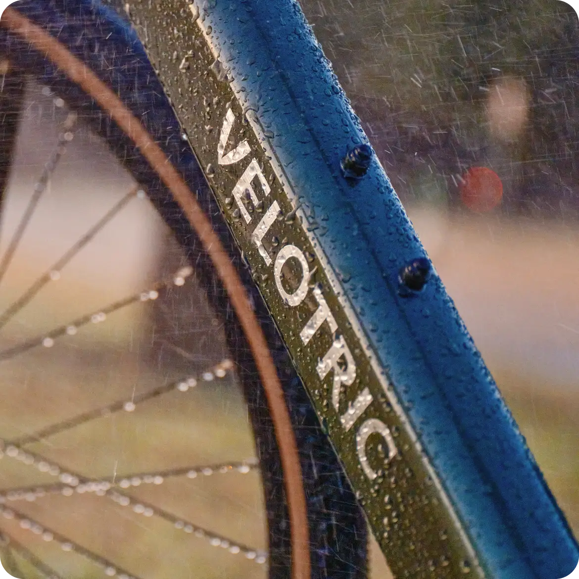 Close-up of the Velotric Tempo by Velotric, showing its blue lightweight city eBike frame and wheel, both covered with raindrops and featuring bold "VELOTRIC" lettering.