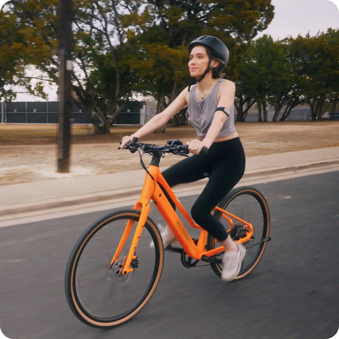 A rider in a helmet cruises on the Velotric Tempo, a lightweight city eBike by Velotric, along a paved road bordered by trees and a fence.