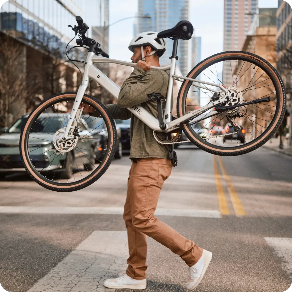 A person wearing a helmet carries the lightweight Velotric Tempo city eBike by Velotric on their shoulder while walking across a city street.