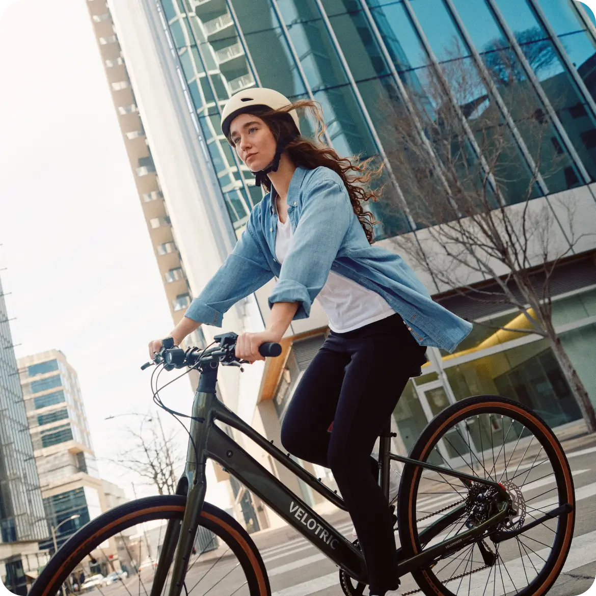 A woman in a helmet rides the Velotric Tempo, a lightweight city eBike by Velotric, along a street with tall glass buildings in the background.